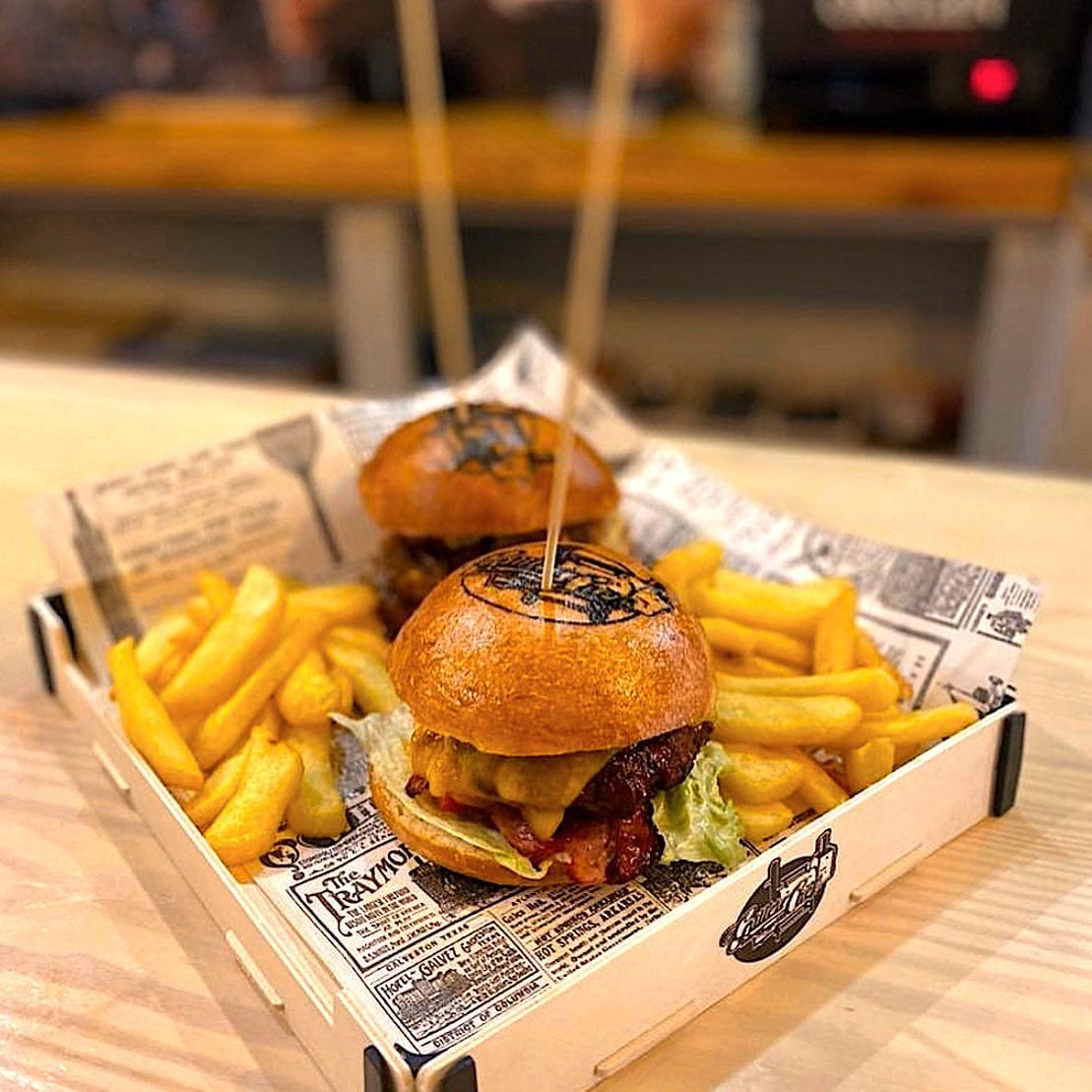 Holztablett mit zwei Burgern und Pommes auf Zeitungspapier, auf einer Bartheke
