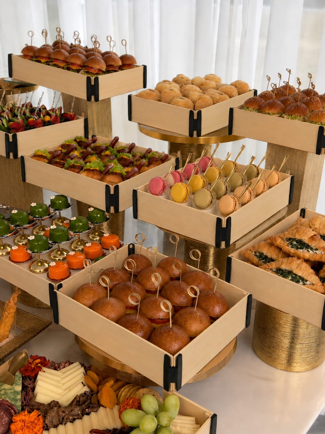 Buffet mit Sincla-Holzkisten ohne Deckel und schwarzen Eckverbindern auf goldenen Ständern, gefüllt mit Mini-Burgern, Brötchen, Macarons, Hotdogs und gefüllten Croissants