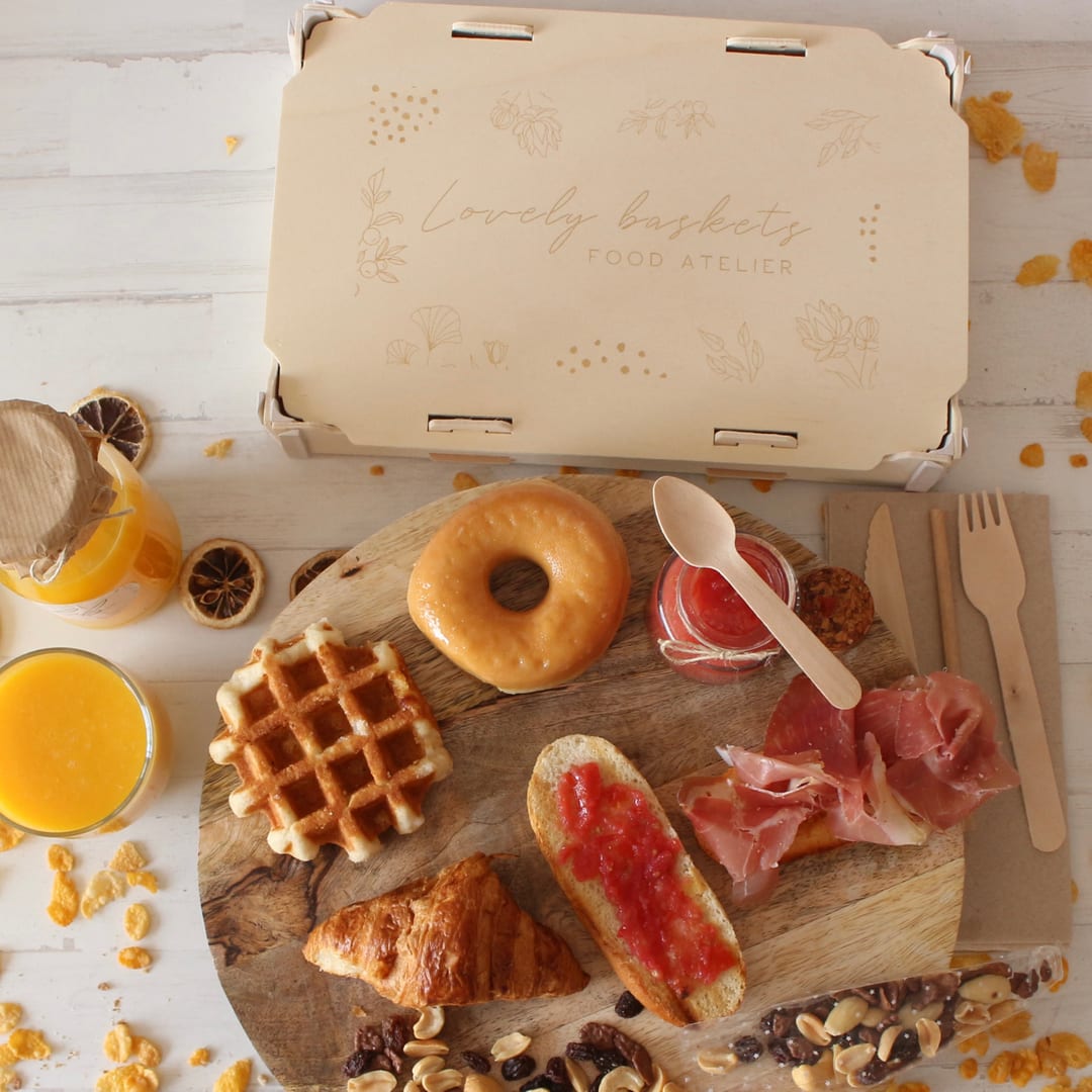 Frühstück auf einem Brett mit Waffel, Donut, Croissant, Brot mit Tomate und Schinken und Orangensaft, vor einem Holzdeckel mit eingraviertem Logo