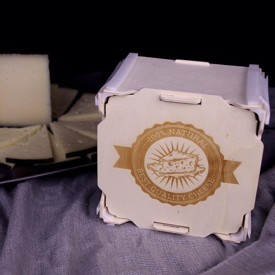 Boîte carrée en bois avec un sceau de fromage gravé sur le couvercle, à côté de morceaux de fromage affiné sur fond sombre