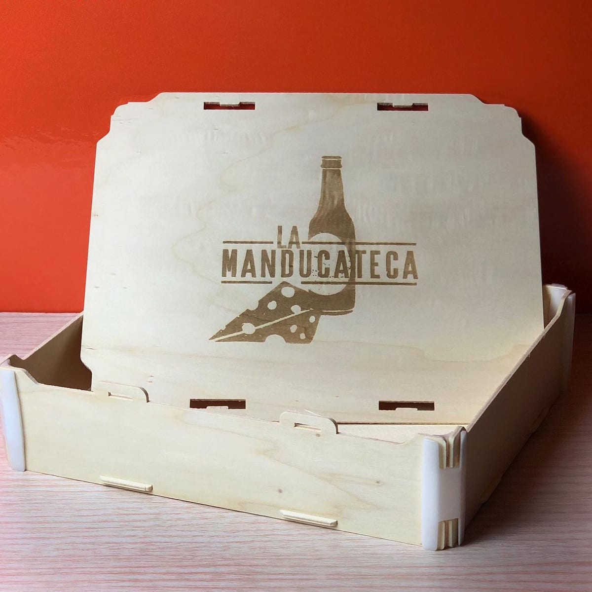 Boîte avec couvercle entrouverte gravée au logo d’une épicerie fine (une bouteille et un morceau de fromage), sur fond orange