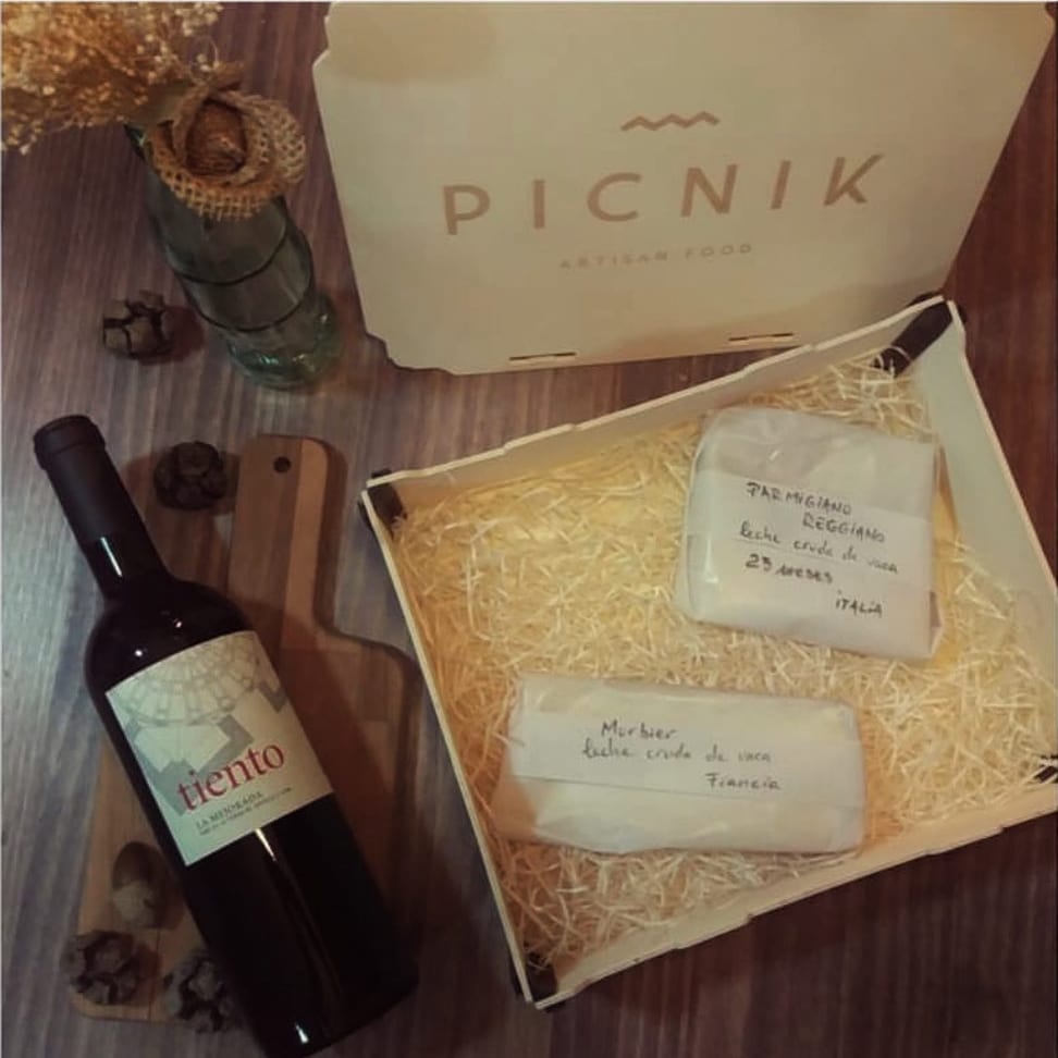 Boîte avec couvercle ouverte avec des fromages emballés dans du papier sur des frisures, à côté d’une bouteille de vin et d’une planche
