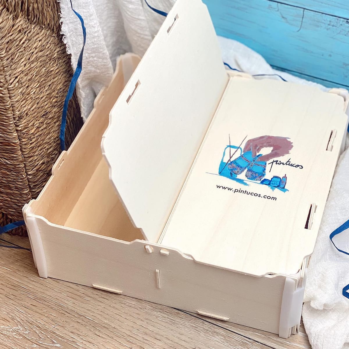 Boîte avec couvercle ouverte, avec à l’intérieur du couvercle le logo illustré en couleur d’un atelier de chaussures pour bébé peintes à la main