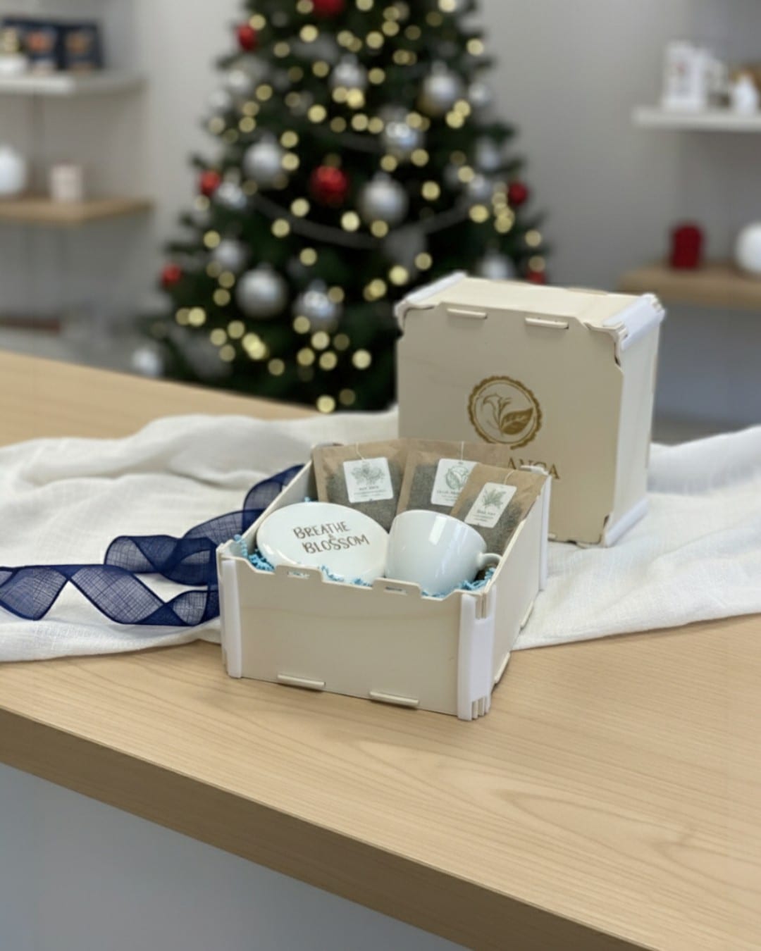 Boîte en bois Sincla avec une tasse, une soucoupe et des sachets de thé, à côté d’une autre boîte au logo gravé, un sapin de Noël en arrière-plan