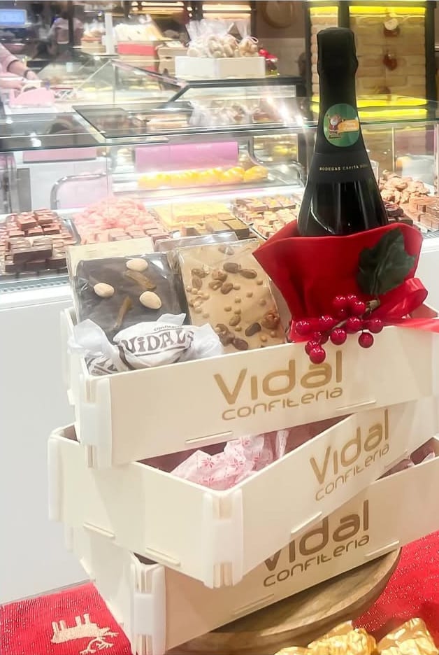 Drei gestapelte Sincla-Holzkisten mit eingraviertem „Vidal confitería“ vorne, mit Turrón und einer Flasche Cava, auf der Theke einer Konditorei