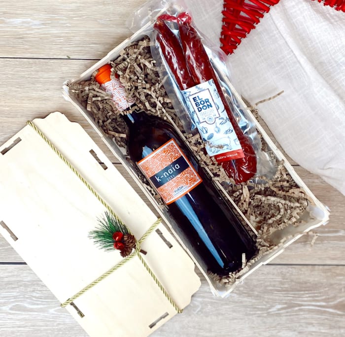 Holzkiste mit Deckel, darin eine Flasche Wein und eine Chorizo auf Papierfüllung