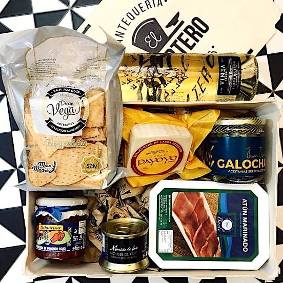 Draufsicht auf eine Holzkiste mit Knabbergebäck, Käse, Oliven, Konfitüre, Pastete und Schinken, neben dem Deckel mit aufgedrucktem Logo eines Feinkostgeschäfts