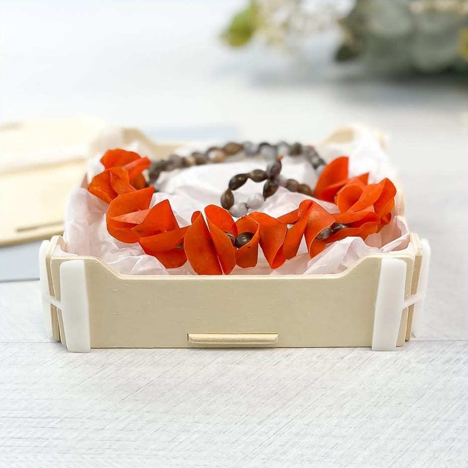 Scatola bassa di legno con una collana artigianale di petali arancioni e semi su carta velina