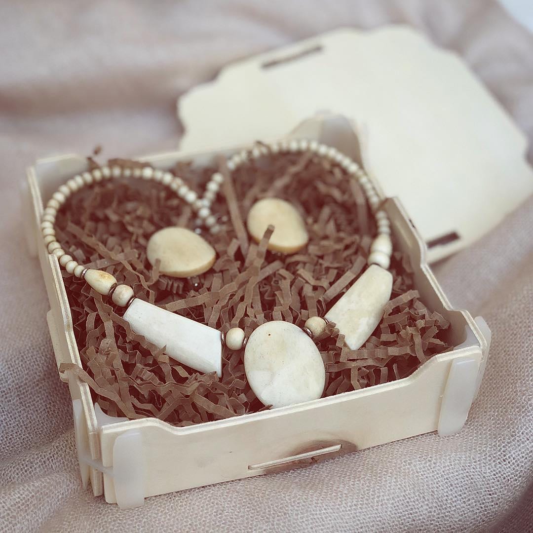 Scatola di legno con trucioli marroni e una collana di perline bianche e pezzi d’osso, con il coperchio appoggiato dietro