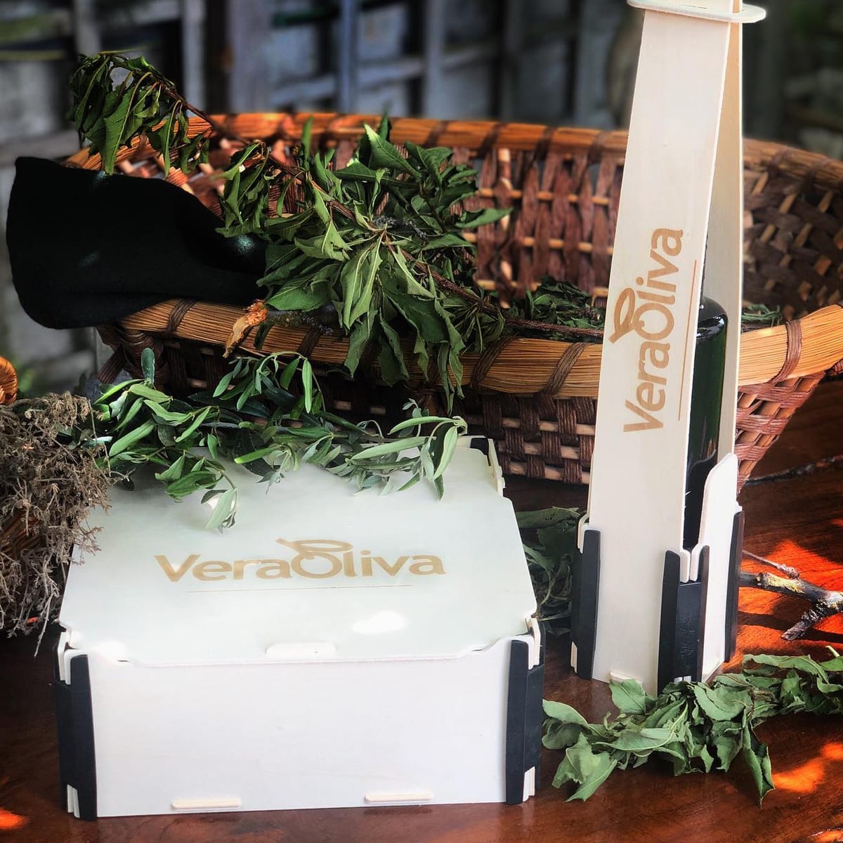 Caja para una botella vertical y caja con tapa con el logo de una marca de aceite grabado, junto a una cesta con ramas y hierbas aromáticas