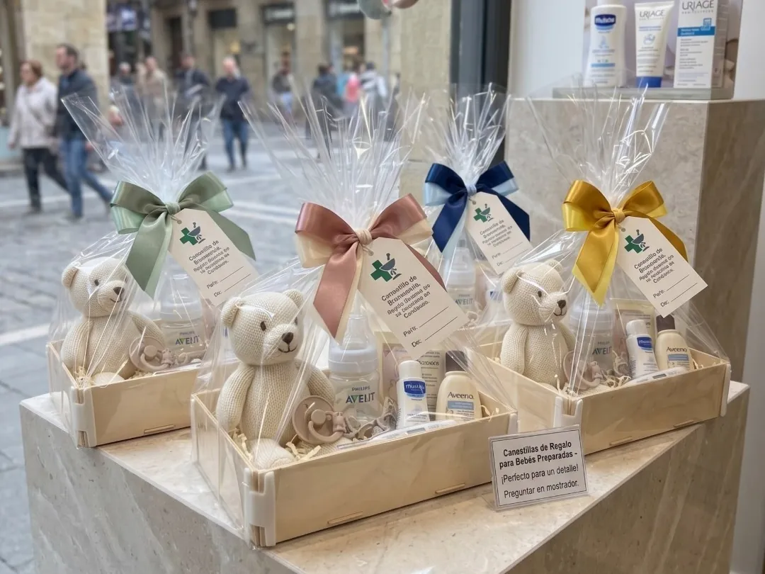 Apothekenschaufenster mit vier Babysets in Holzkisten, mit Teddybären und bunten Schleifen, dahinter die Straße
