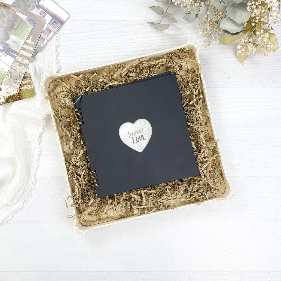 Caixa de madeira Sincla com um álbum de fotos preto sobre papel kraft frisado; ao lado, fotos de casamento impressas, uma gaze branca e flores secas