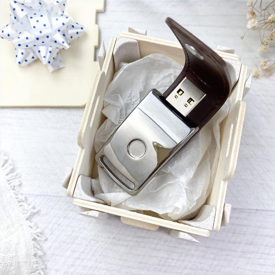 Caixa pequena aberta com uma pen USB de pele castanha sobre papel de seda, com a tampa e um laço ao lado
