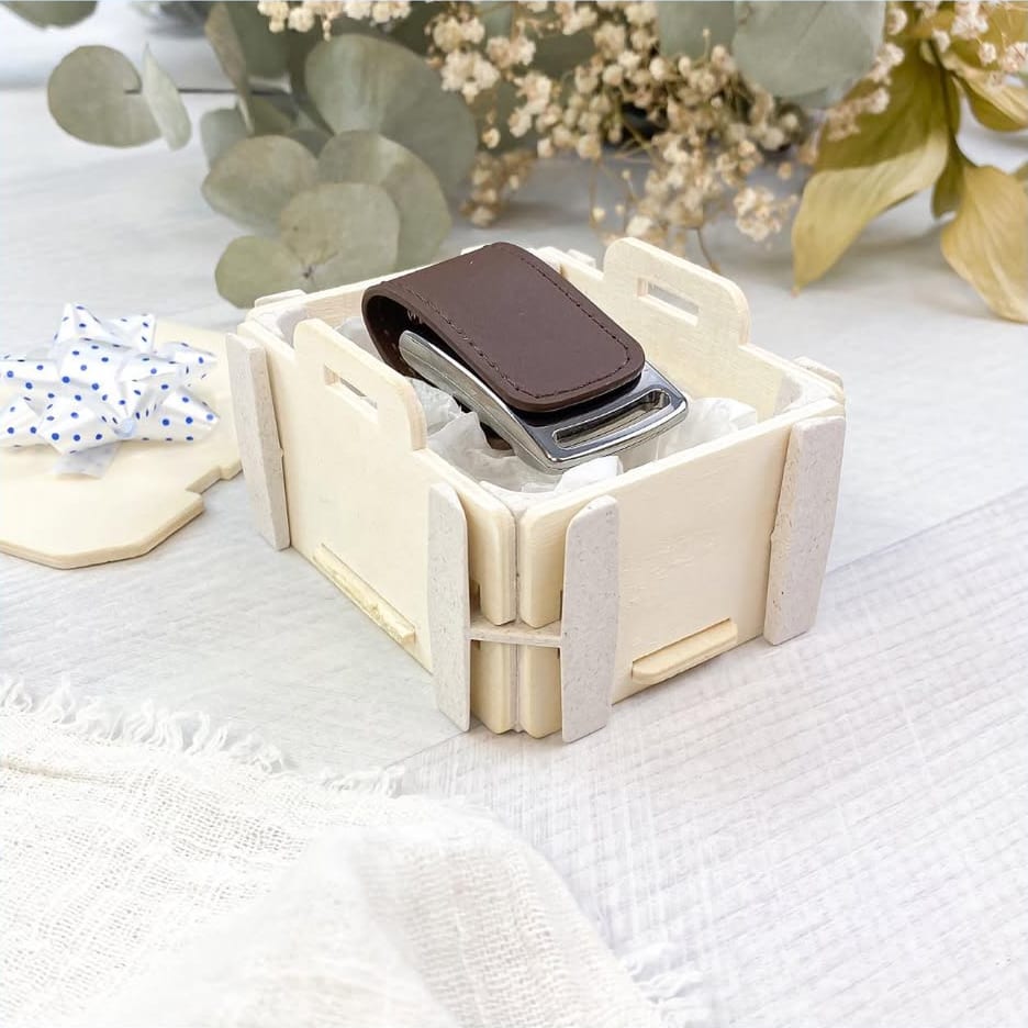 Caixa pequena de madeira Sincla com uma pen USB de pele castanha sobre papel de seda; ao lado, a tampa com um laço às bolinhas, eucalipto e flores secas