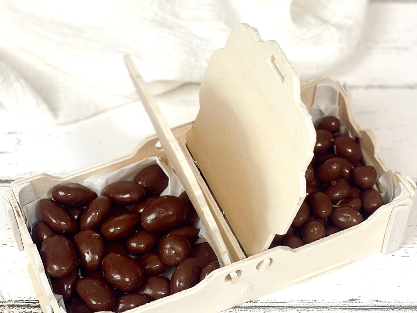Cesta de madera de dos compartimentos con la tapa levantada y almendras cubiertas de chocolate a cada lado del separador