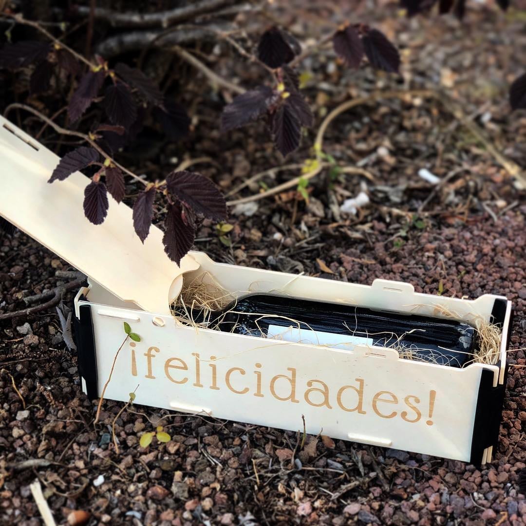 Wooden box for a bottle lying flat, with “¡felicidades!” engraved on the front and the lid half open