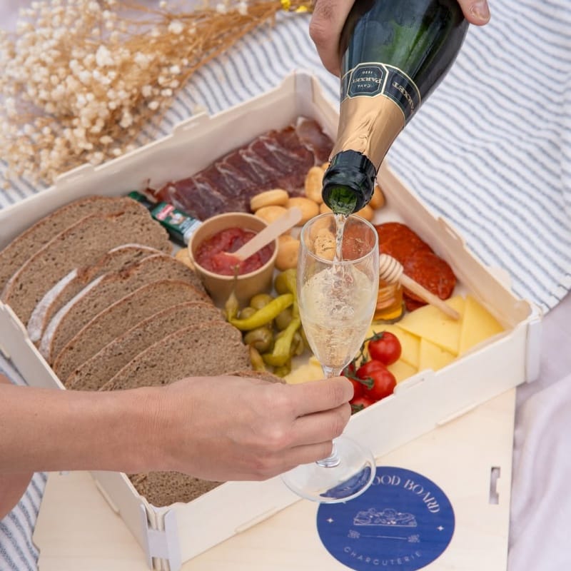 Person pouring cava into a glass above a wooden box filled with bread, charcuterie, cheese and pickles
